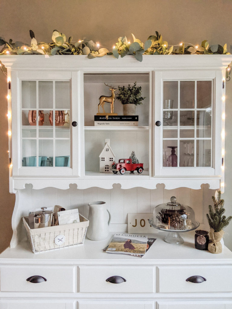 white dining hutch filled with christmas decor, mugs, glassware, garland and twinkle lights