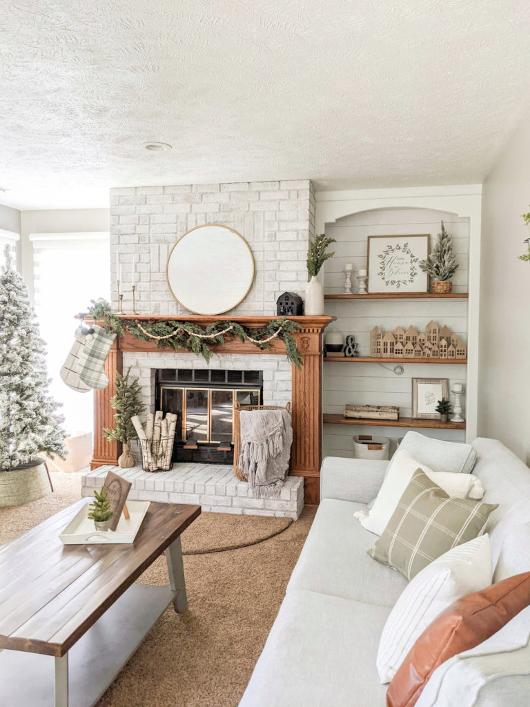 christmas decorated living room with a brick fireplace, grey couch, and self inserts decorated with trees, garland, wooden houses, wood, pillows and blankets