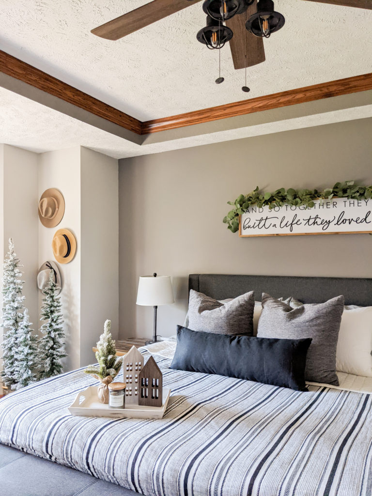 king sized bed with striped bedding, grey and black pillows, and a white tray decorated with wooden houses, a small christmas tree and a candle