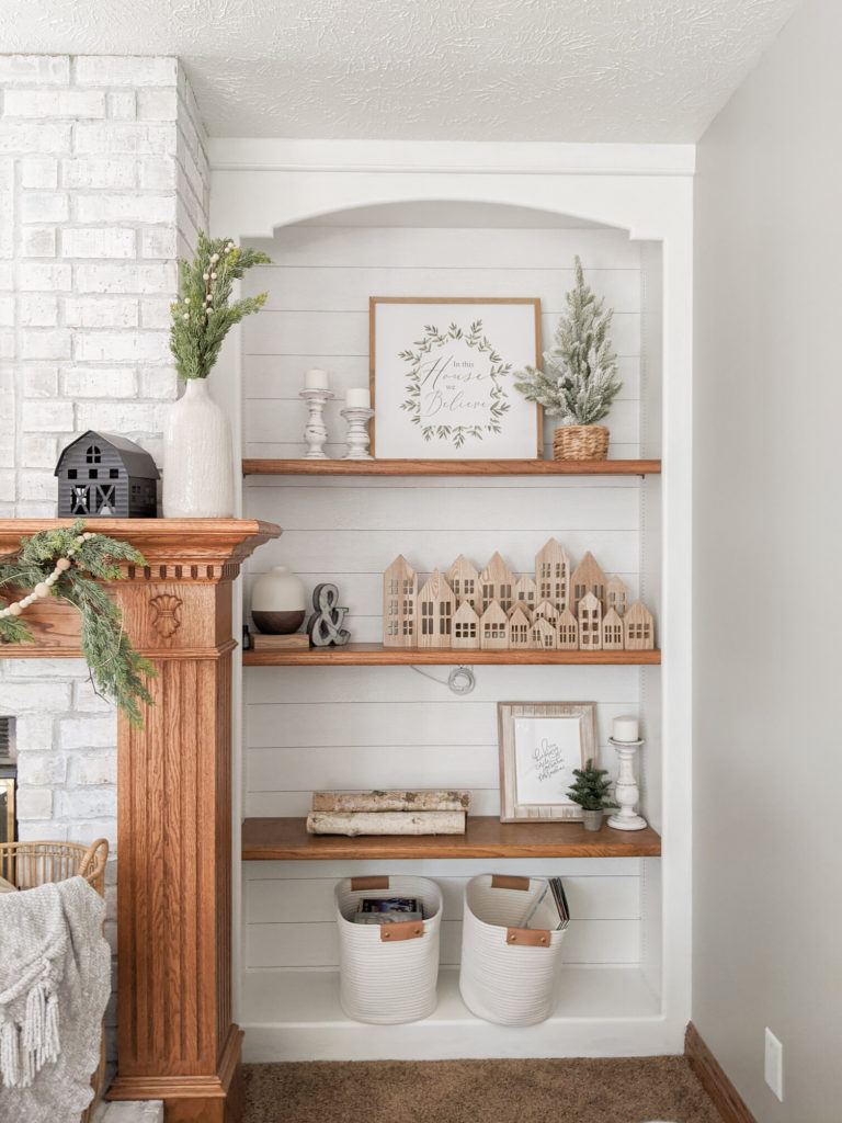 a christmas decorated shelf with a wooden village, sign, candles, trees, and baskets