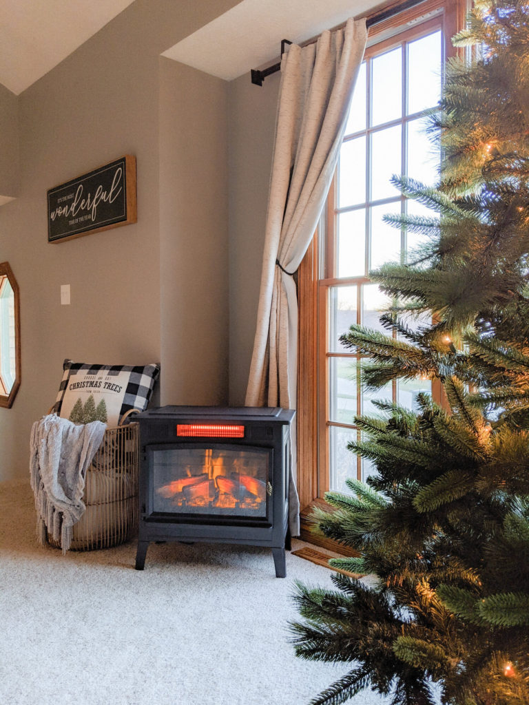 an electric fireplace stove tucked in the corner of a small living room with a basket of pillows and a lit up Christmas tree next to it